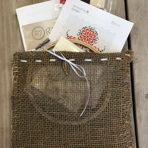 May include: A cross-stitch kit with a burlap bag. The kit includes a pattern labeled "The Tree Of Life", embroidery floss in red, brown, and cream, a needle, and a wooden embroidery hoop. The bag has a leaf-shaped charm.