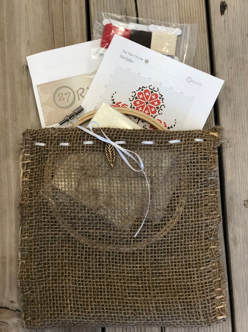 May include: A cross-stitch kit with a burlap bag. The kit includes a pattern labeled "The Tree Of Life", embroidery floss in red, brown, and cream, a needle, and a wooden embroidery hoop. The bag has a leaf-shaped charm.