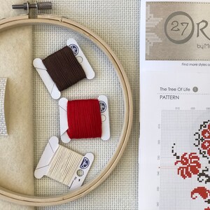 May include: Cross-stitch supplies including a wooden embroidery hoop, three spools of thread in brown, red, and cream, and a piece of fabric with a needle. A pattern sheet with the text "The Tree Of Life" is also visible.