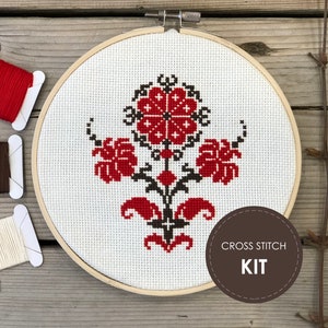 May include: A cross stitch kit featuring a floral design in red and black on white fabric, mounted in a wooden hoop. The kit includes red, brown, and cream-colored thread spools. The text "CROSS STITCH KIT" is on a brown circle.