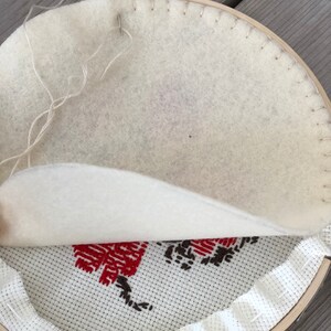 May include: A cross-stitch project in progress, featuring a wooden embroidery hoop. The fabric displays a red and brown floral design. A needle is inserted through the white backing fabric, with loose white thread visible.