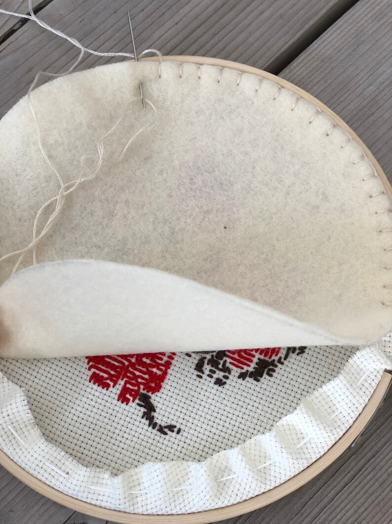 May include: A cross-stitch project in progress, featuring a wooden embroidery hoop. The fabric displays a red and brown floral design. A needle is inserted through the white backing fabric, with loose white thread visible.