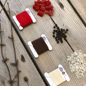 May include: An overhead shot of a wooden surface with three spools of DMC embroidery floss in red, brown, and cream colors. Scattered around are red diced peppers, black peppercorns, and white rice. Dried twigs are on the left.