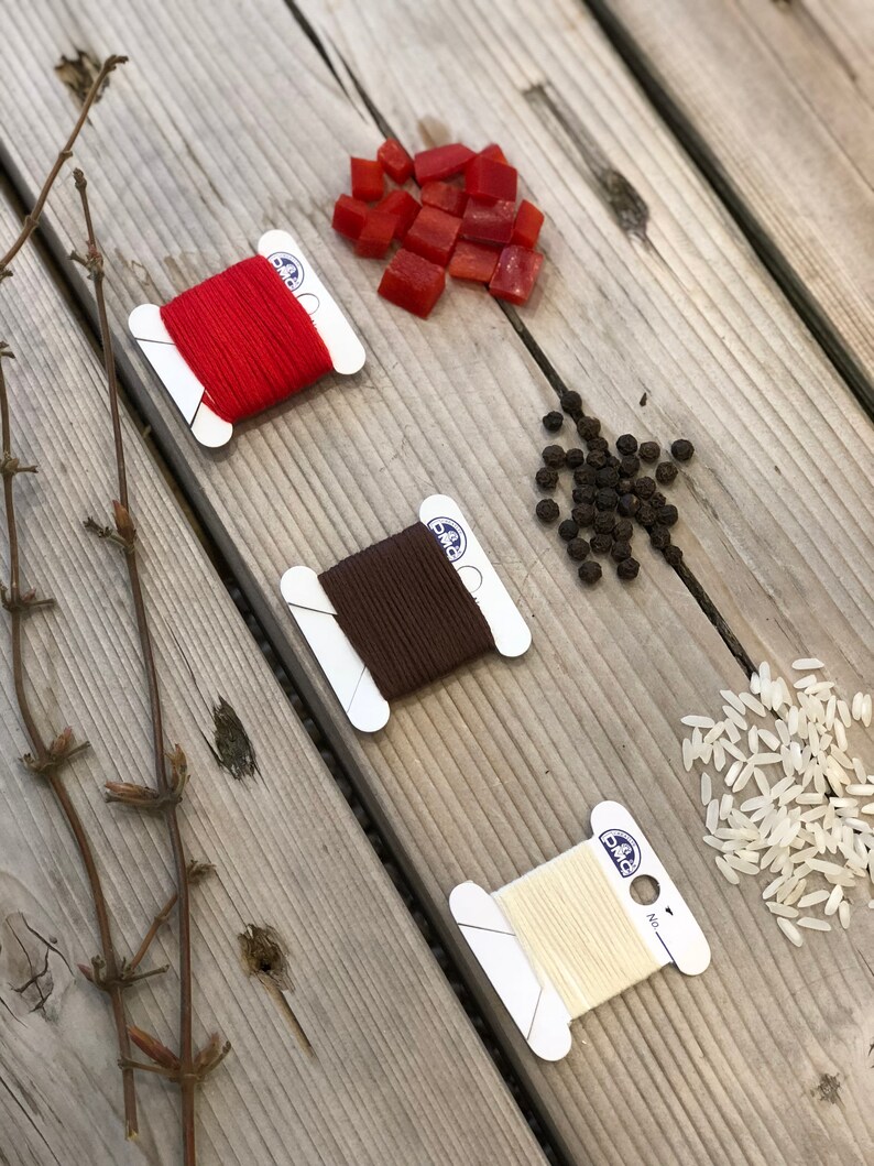 May include: An overhead shot of a wooden surface with three spools of DMC embroidery floss in red, brown, and cream colors. Scattered around are red diced peppers, black peppercorns, and white rice. Dried twigs are on the left.