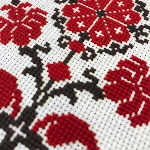 May include: Close-up of a cross-stitch floral design. The embroidery features red flowers and leaves with black stems and accents on a white background. The stitches are visible, creating a textured effect.