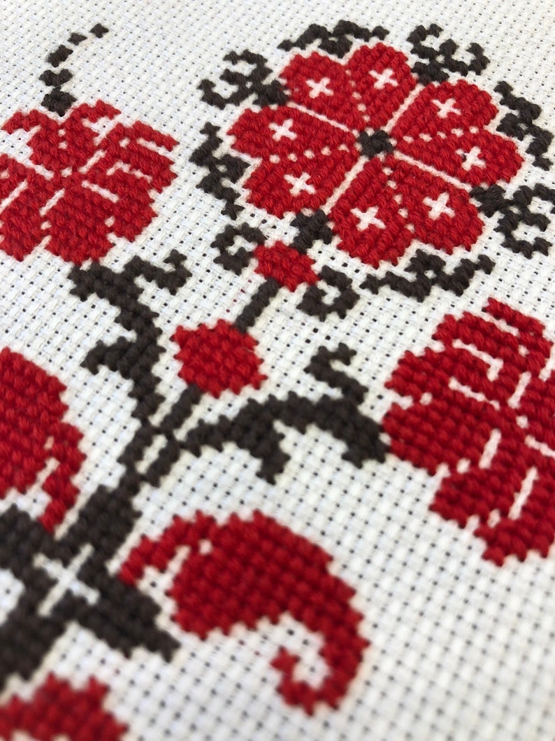 May include: Close-up of a cross-stitch floral design. The embroidery features red flowers and leaves with black stems and accents on a white background. The stitches are visible, creating a textured effect.