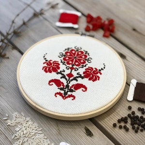 May include: A completed cross-stitch embroidery in a wooden hoop. The design features a red and black floral pattern on a white background. Spools of thread, rice, and peppercorns are scattered around the hoop on a wooden surface.