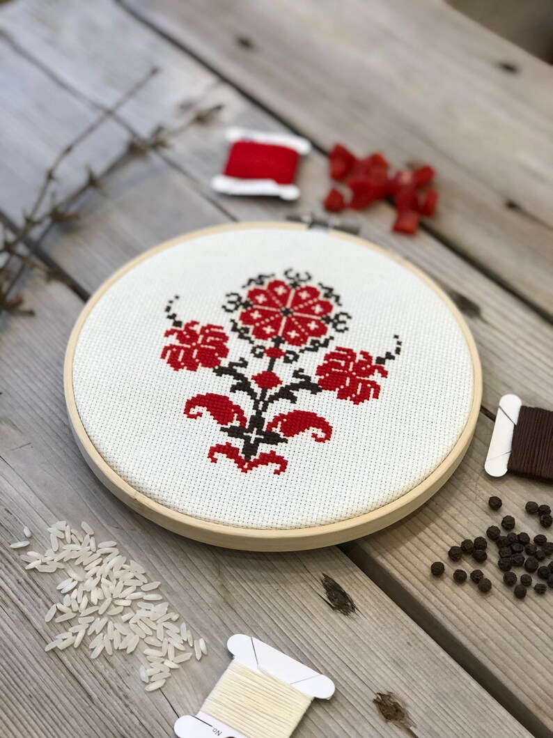 May include: A completed cross-stitch embroidery in a wooden hoop. The design features a red and black floral pattern on a white background. Spools of thread, rice, and peppercorns are scattered around the hoop on a wooden surface.