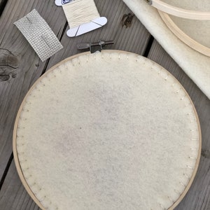 May include: A craft supplies set featuring a wooden embroidery hoop with cream-colored fabric, a spool of off-white thread, a needle, and a piece of fabric with a grid pattern. The items are arranged on a weathered wooden surface, suggesting a DIY or crafting theme.