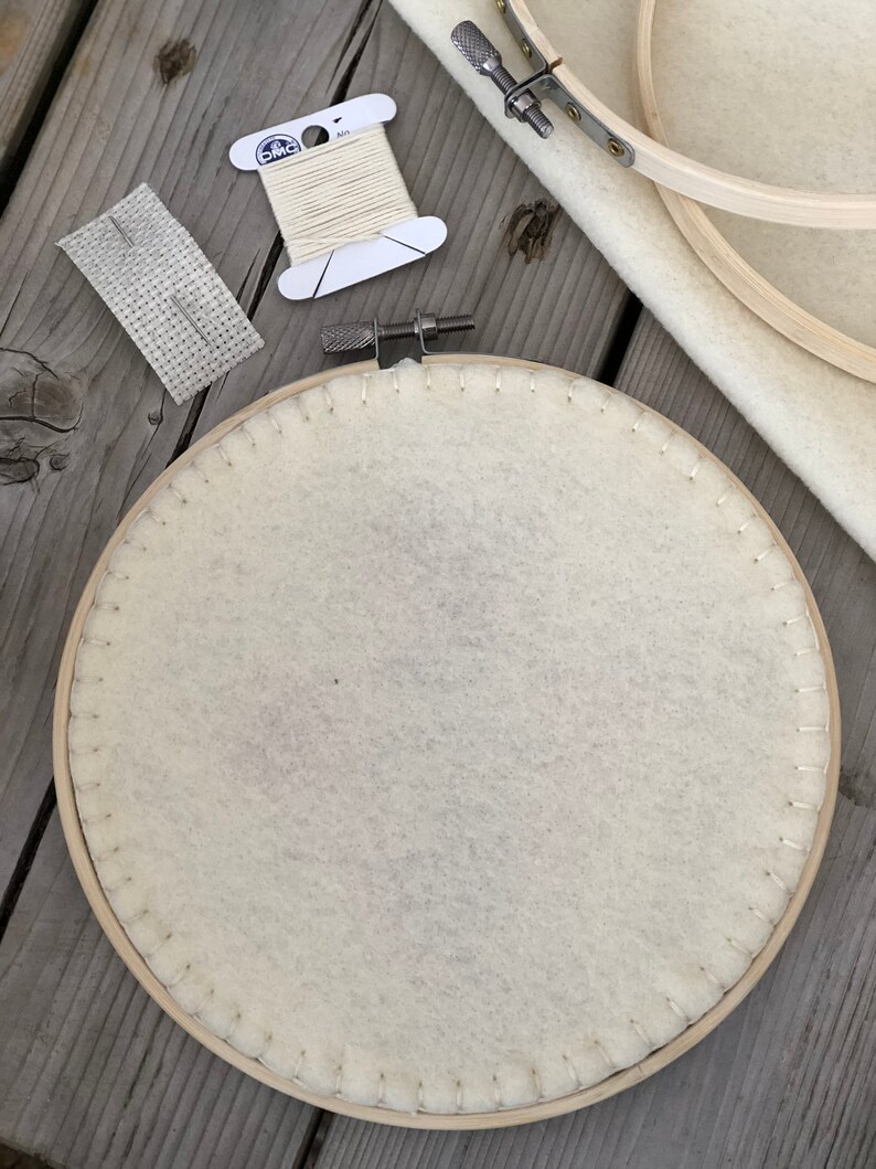 May include: A craft supplies set featuring a wooden embroidery hoop with cream-colored fabric, a spool of off-white thread, a needle, and a piece of fabric with a grid pattern. The items are arranged on a weathered wooden surface, suggesting a DIY or crafting theme.