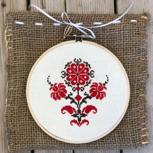 May include: A round, white cross-stitch embroidery hoop featuring a floral design in red and black. The embroidery is mounted on a burlap backing and tied with a white ribbon and a wooden leaf accent. The piece is displayed on a wooden surface.