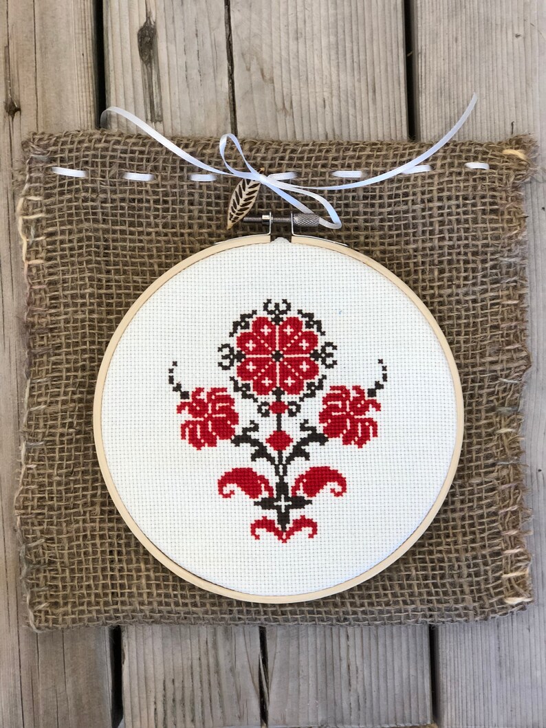 May include: A round, white cross-stitch embroidery hoop featuring a floral design in red and black. The embroidery is mounted on a burlap backing and tied with a white ribbon and a wooden leaf accent. The piece is displayed on a wooden surface.