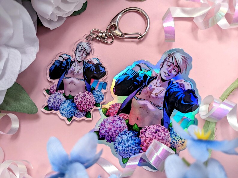 May include: Two clear acrylic charms featuring a shirtless figure with a blue jacket, surrounded by purple, pink, and blue flowers. One charm has a silver keychain attached. The background is pink with floral accents.