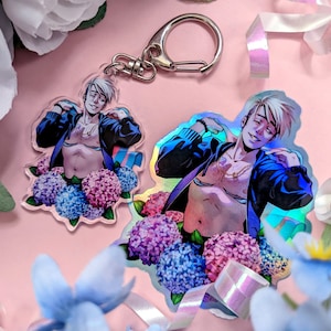 May include: Two clear acrylic charms featuring a smiling person with a black jacket and a floral arrangement of blue and pink hydrangeas. One charm is a keychain, the other is a sticker with a holographic effect.