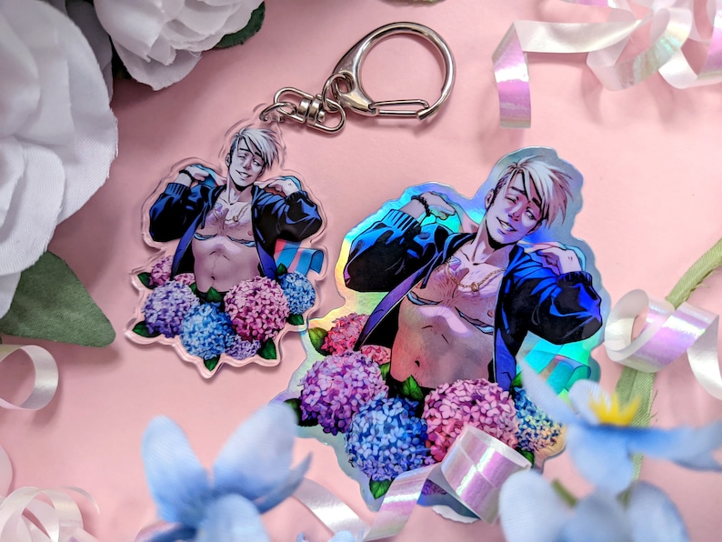 May include: Two clear acrylic charms featuring a smiling person with a black jacket and a floral arrangement of blue and pink hydrangeas. One charm is a keychain, the other is a sticker with a holographic effect.
