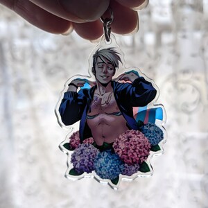 May include: A clear acrylic keychain featuring a stylized illustration of a person with a blue jacket, surrounded by blue and pink flowers. The person is shirtless and has a necklace. A transgender flag is also visible.
