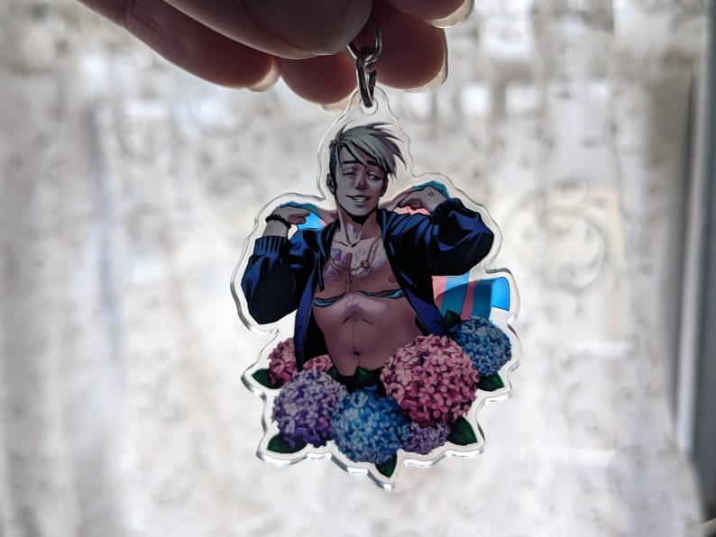 May include: A clear acrylic keychain featuring a stylized illustration of a person with a blue jacket, surrounded by blue and pink flowers. The person is shirtless and has a necklace. A transgender flag is also visible.