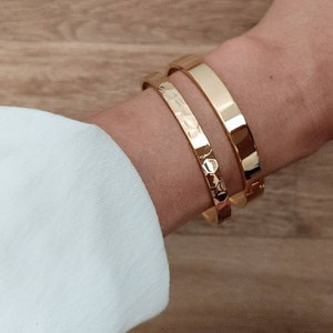May include: Two gold bracelets on a person's wrist. The bracelet on top is smooth and the bracelet on the bottom has a textured design.