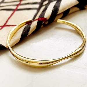 Może przedstawiać: Złota bransoletka typu bangle o gładkim, zakrzywionym designie. Bransoletka jest otwarta i ma prosty, elegancki styl.