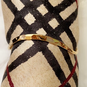 May include: A gold-toned hammered metal bangle bracelet. The bracelet is slightly curved and has a textured surface.