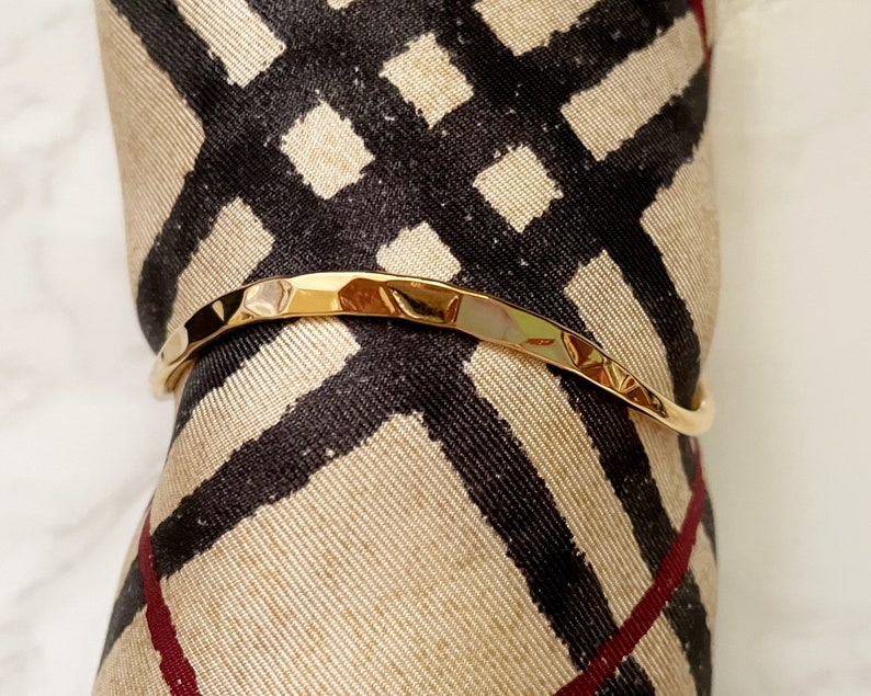 May include: A gold-toned hammered metal bangle bracelet. The bracelet is slightly curved and has a textured surface.