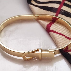 Könnte beinhalten: Ein goldfarbenes Armreif-Armband mit einer Schließe. Das Armband ist schlicht und elegant.