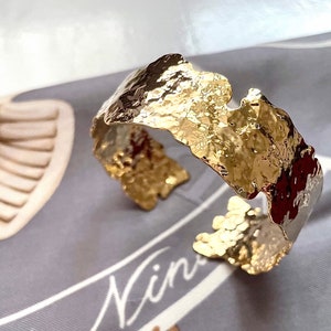 Può includere: Un braccialetto rigido in metallo martellato color oro con una superficie irregolare e testurizzata. Il braccialetto è aperto e ha una forma leggermente curva.