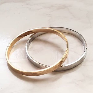 May include: Two metal bangles, one gold and one silver, are stacked on top of each other. Both bangles have a hammered texture.