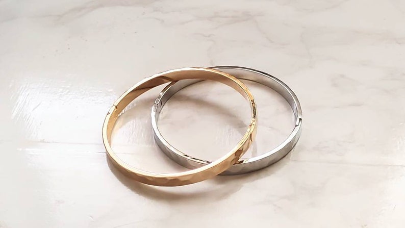 May include: Two metal bangles, one gold and one silver, are stacked on top of each other. Both bangles have a hammered texture.