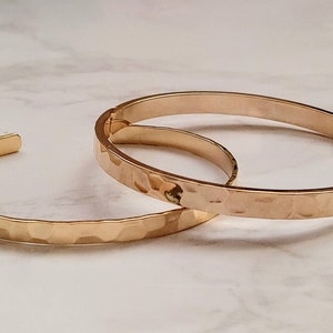 May include: Two gold-toned hammered metal bangle bracelets. The bracelets are stacked on top of each other.