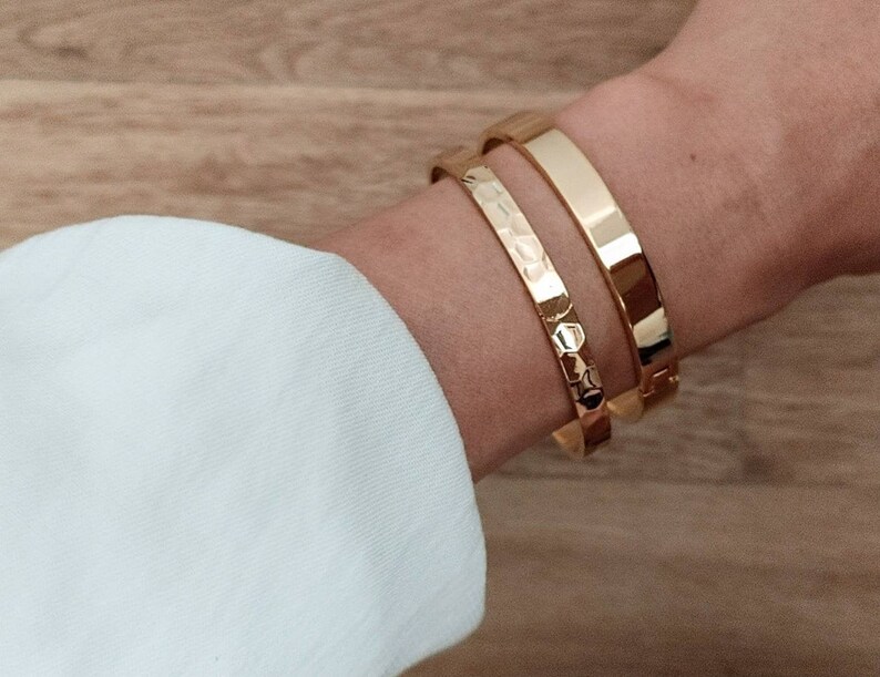 May include: Two gold bracelets on a person's wrist. The bracelet on top is smooth and the bracelet on the bottom has a textured design.