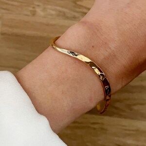 May include: A gold-toned hammered metal bangle bracelet. The bracelet is open and has a slightly curved shape.