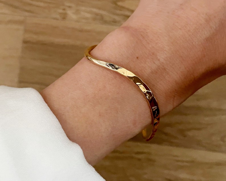 May include: A gold-toned hammered metal bangle bracelet. The bracelet is open and has a slightly curved shape.