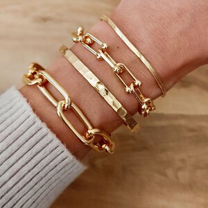 May include: Four gold bracelets stacked on a wrist. The bottom bracelet is a thick, chunky chain link style. The next bracelet is a hammered gold band. The third bracelet is a delicate chain with small round accents. The top bracelet is a thin, shiny, snake chain.