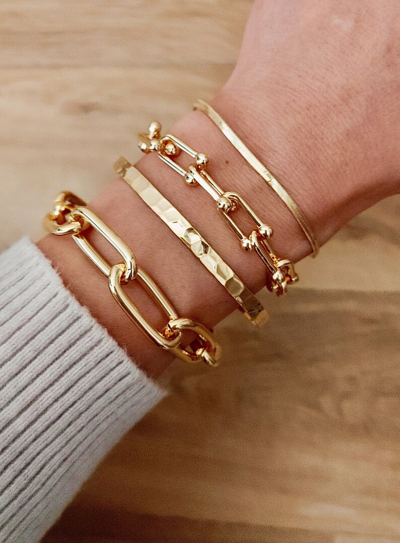 May include: Four gold bracelets stacked on a wrist. The bottom bracelet is a thick, chunky chain link style. The next bracelet is a hammered gold band. The third bracelet is a delicate chain with small round accents. The top bracelet is a thin, shiny, snake chain.