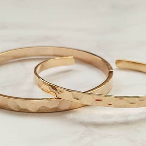 May include: Two gold hammered metal bangles, one larger than the other, are stacked on a white surface.