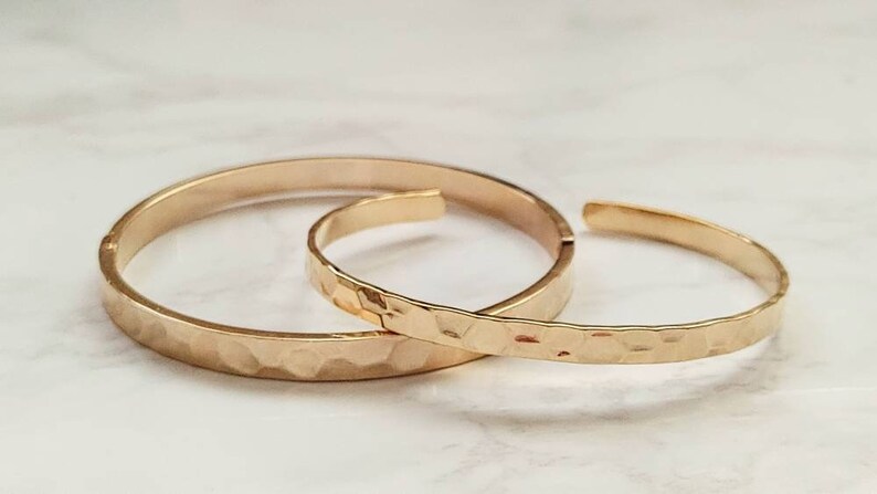 May include: Two gold hammered metal bangles, one larger than the other, are stacked on a white surface.