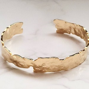 May include: A gold-toned, textured metal cuff bracelet with a hammered finish. The bracelet is open and has a slightly irregular shape.