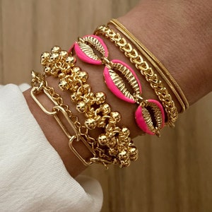 May include: A set of gold bracelets with pink shell charms. The bracelets are layered on a person's wrist.