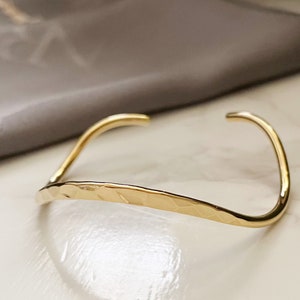 May include: A gold-toned hammered metal bangle bracelet. The bracelet is open and has a slightly curved shape.
