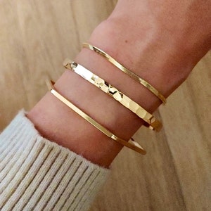 May include: Three gold bracelets stacked on a wrist. The top bracelet is a thin, shiny, snake chain. The middle bracelet is a hammered, textured band. The bottom bracelet is a thick, flat band.