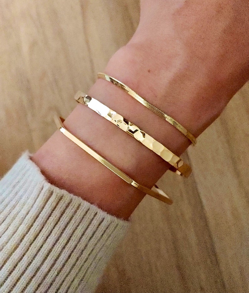 May include: Three gold bracelets stacked on a wrist. The top bracelet is a thin, shiny, snake chain. The middle bracelet is a hammered, textured band. The bottom bracelet is a thick, flat band.