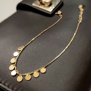 以下が含まれることがあります： 小さな丸いチャームがぶら下がった金のチェーンネックレス。ネックレスは黒い革の表面の上に置かれています。