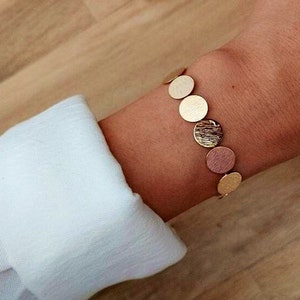 Puede incluir: Pulsera dorada con una cadena de discos metálicos redondos y texturizados.