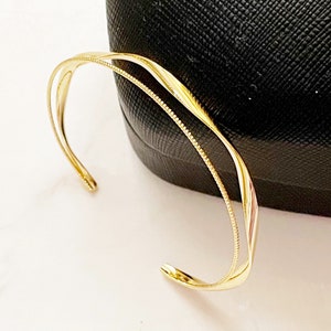 May include: A gold-colored open cuff bracelet with a twisted design. The bracelet has a smooth, polished finish and a delicate, beaded detail along one edge. The bracelet is resting on a black surface.