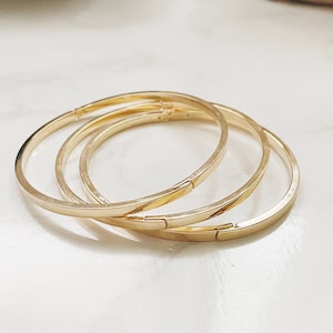 May include: Three gold-colored bangles are displayed on a white marble surface. The bracelets have a simple, elegant design with a flat profile and a clasp closure. The bangles are stacked together, creating a layered look.