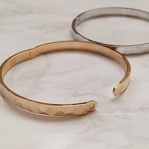 May include: Two simple bangle bracelets, one gold and one silver, with a hammered texture. The gold bracelet is open and the silver bracelet is closed.