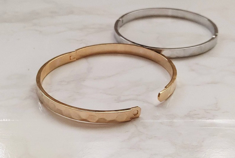May include: Two simple bangle bracelets, one gold and one silver, with a hammered texture. The gold bracelet is open and the silver bracelet is closed.