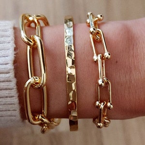 May include: Three gold bracelets stacked on a wrist. The top bracelet is a delicate chain with small round accents. The middle bracelet is a thick, hammered band. The bottom bracelet is a chunky chain with large oval links.