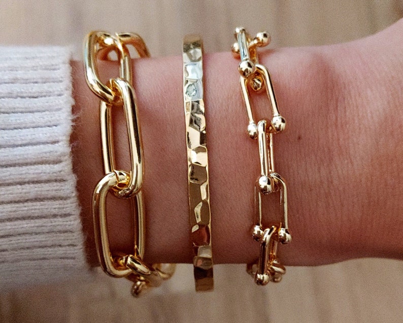 May include: Three gold bracelets stacked on a wrist. The top bracelet is a delicate chain with small round accents. The middle bracelet is a thick, hammered band. The bottom bracelet is a chunky chain with large oval links.